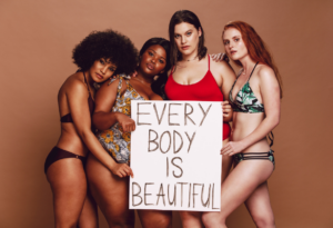 Vier Frauen in Bademode halten ein Schild mit der Aufschrift ‚Every Body Is Beautiful‘.