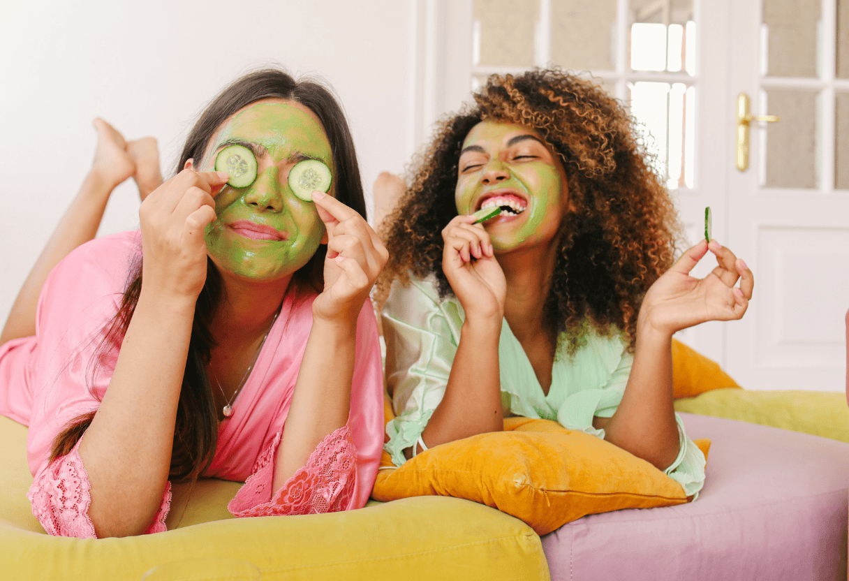Zwei Frauen mit Gesichtsmasken und Gurkenscheiben lachen während eines Spa-Tags zu Hause