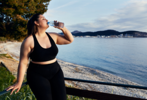 Frau in Sportkleidung trinkt Wasser am Strand nach einem Training