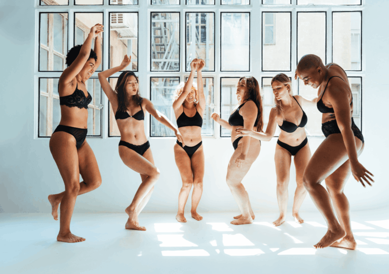 Sechs Body Neutrality Frauen unterschiedlicher Körperformen tanzen in schwarzer Unterwäsche vor einem Fenster
