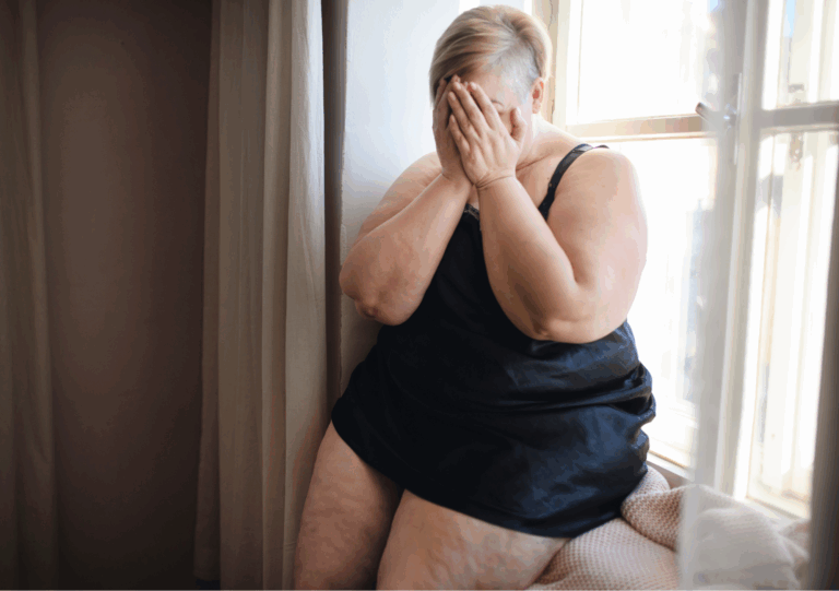Traurige Frau sitzt am Fenster und hält sich das Gesicht vor Scham und innerem Schmerz