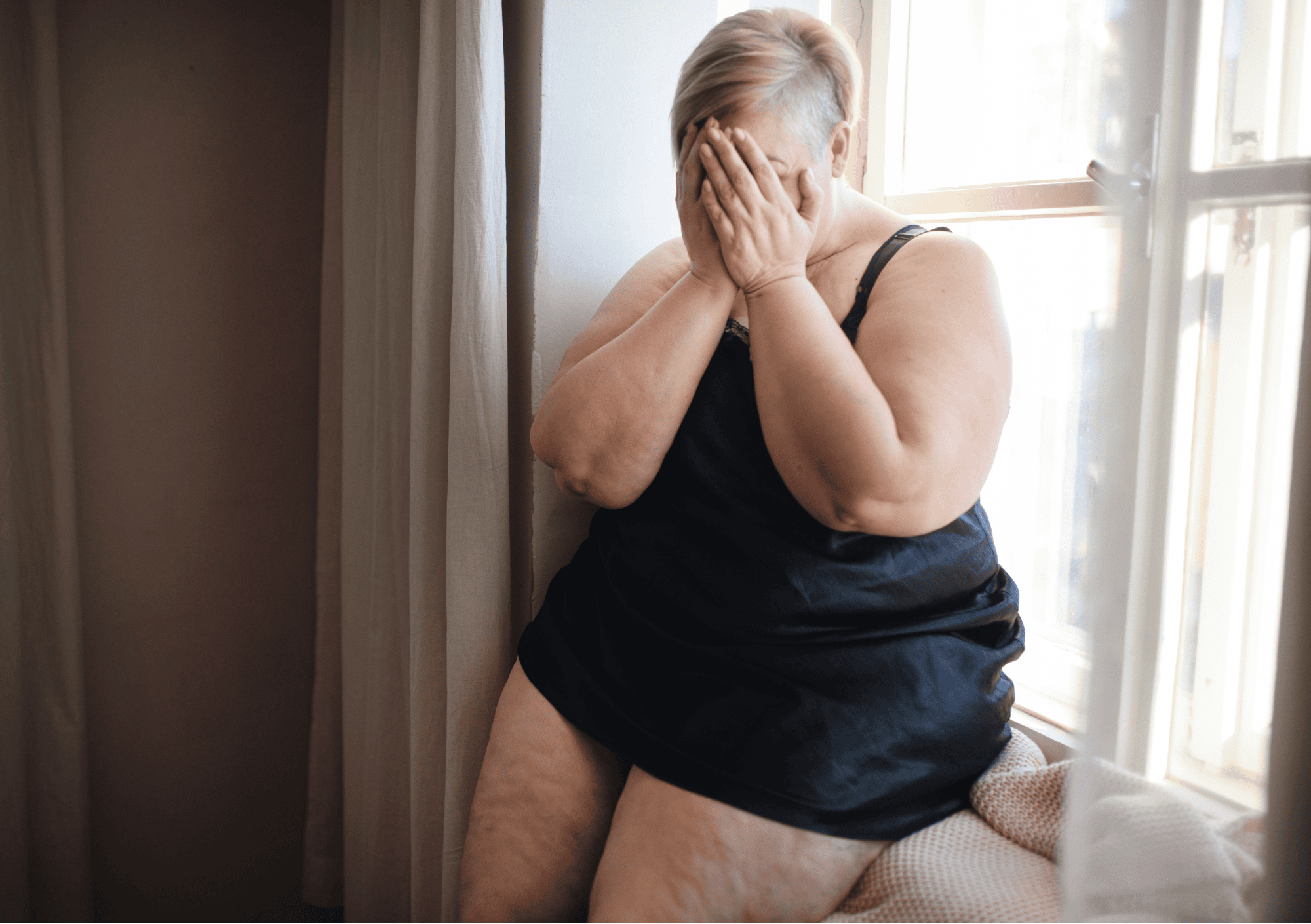 Traurige Frau sitzt am Fenster und hält sich das Gesicht vor Scham und innerem Schmerz