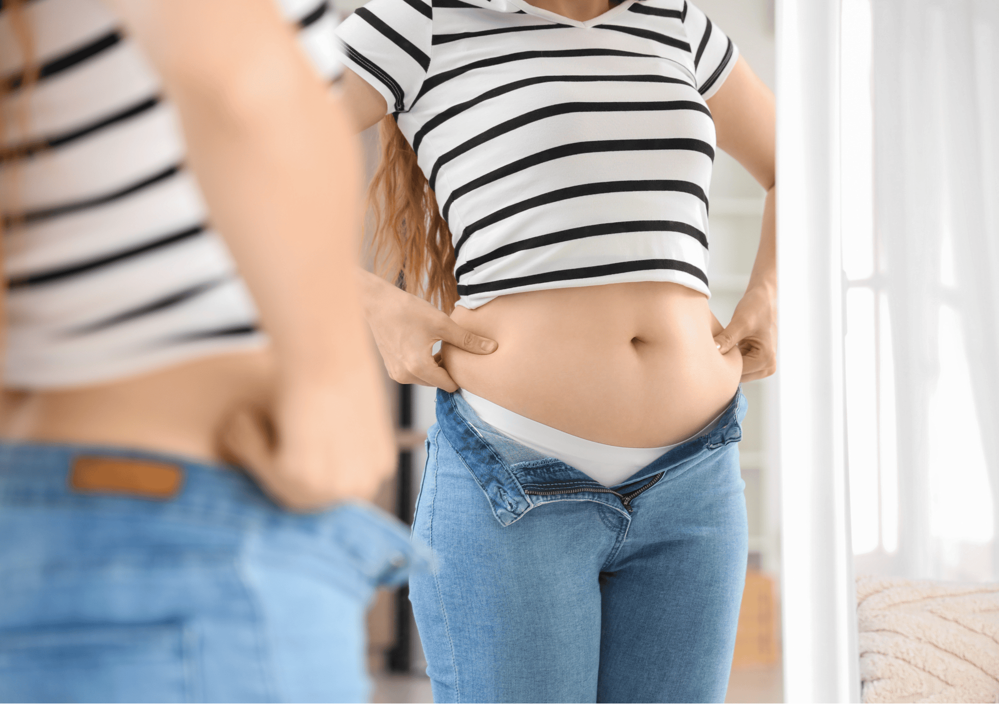 Junge Frau steht vor dem Spiegel und findet ihren Bauch zu fett.