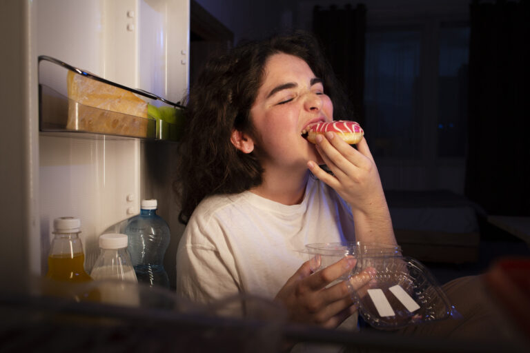 Junge Frau isst spät abends emotional einen Donut vor offenem Kühlschrank