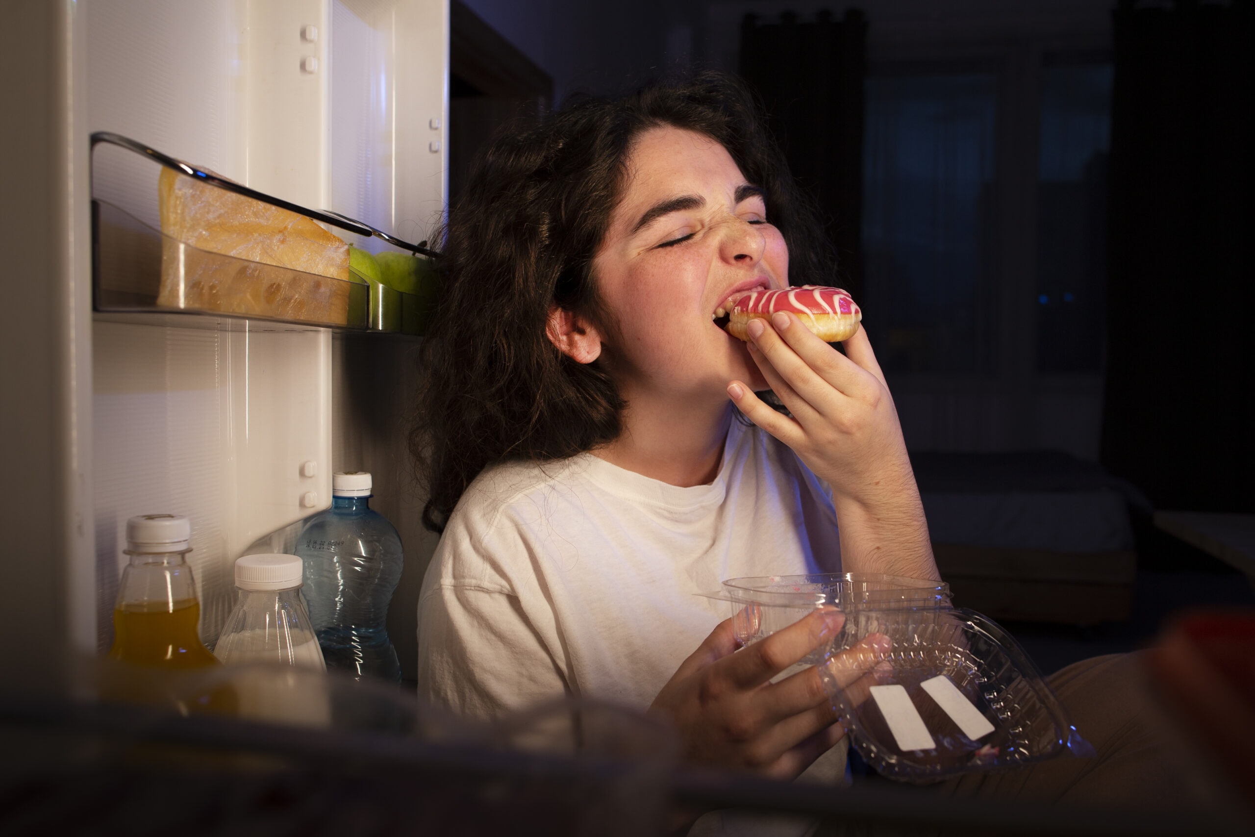 Junge Frau isst spät abends emotional einen Donut vor offenem Kühlschrank