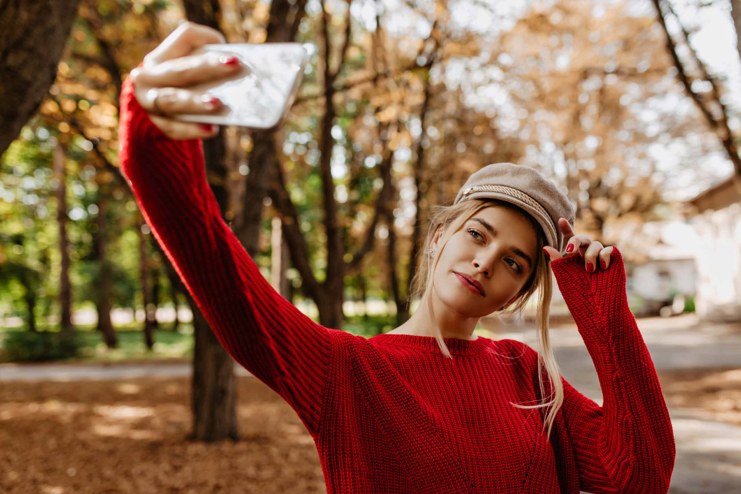 Frau im roten Pullover und Mütze macht Selfie im herbstlichen Park