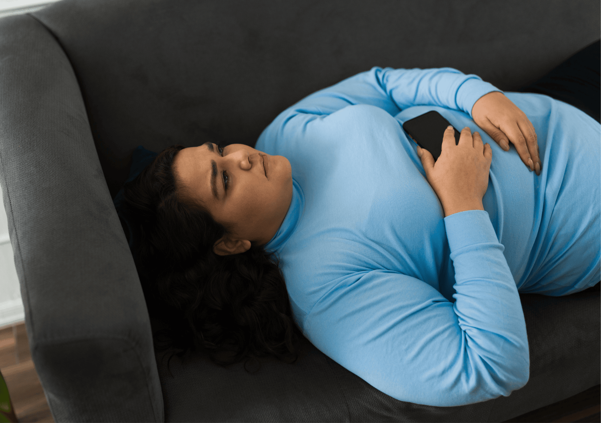 Frau in blauem Pullover liegt nachdenklich mit Handy auf dem Sofa