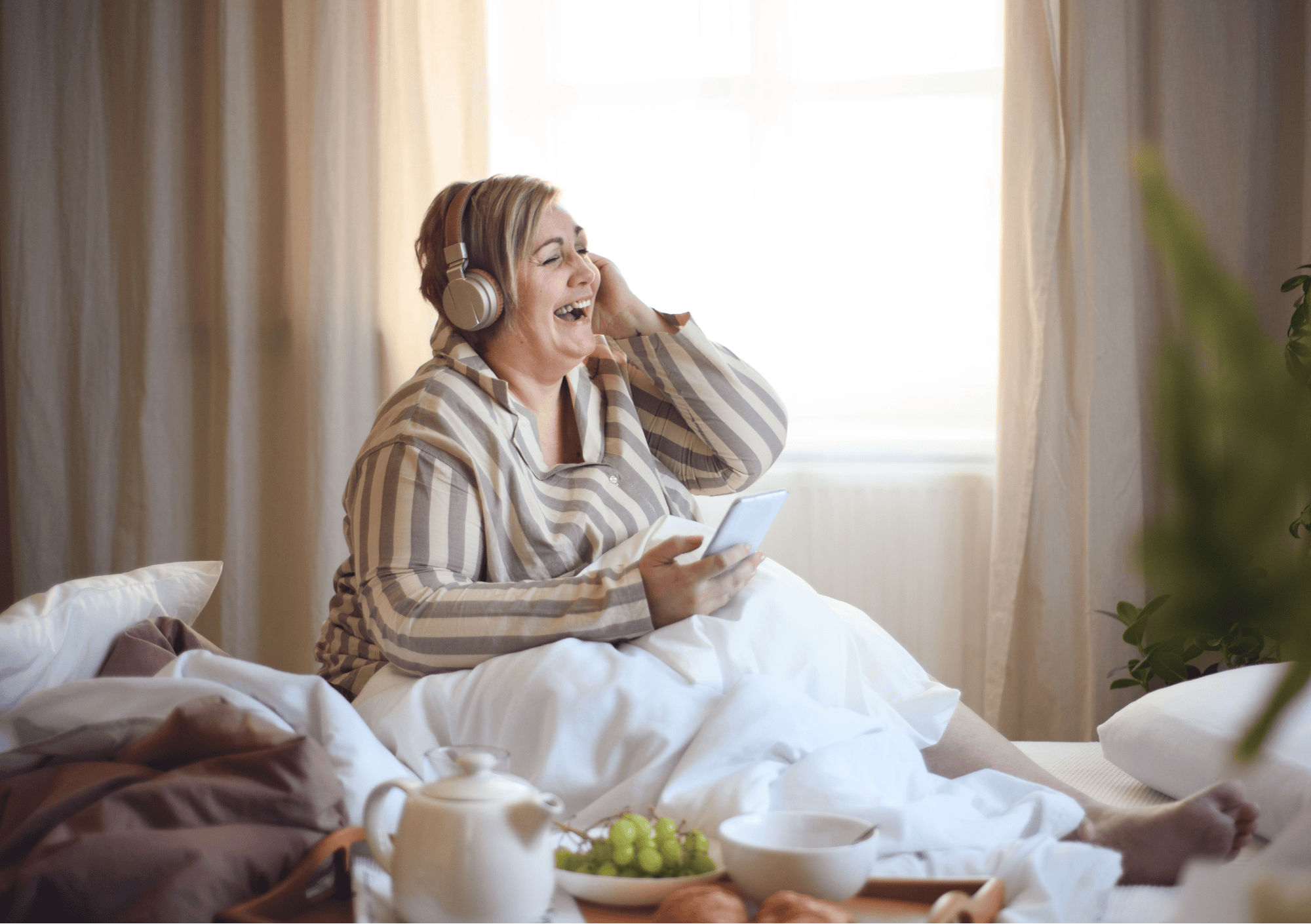 Lachende Frau im Bett hört Podcast mit Frühstückstablett – Moment der Selbstfürsorge