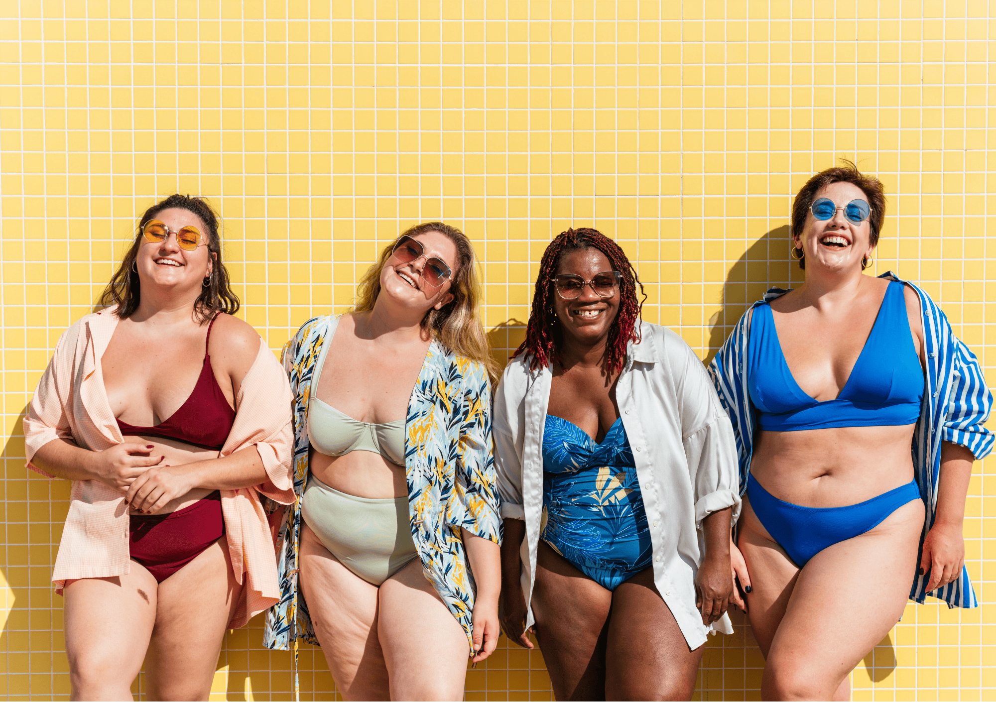 Diverse Frauengruppe in Bikinis und Badeanzügen lacht vor einer gelben Kachelwand