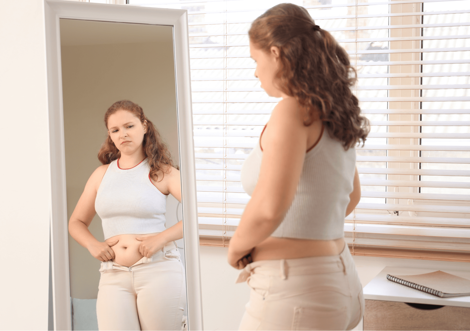Junge Frau schaut kritisch in den Spiegel und betrachtet ihren Bauch