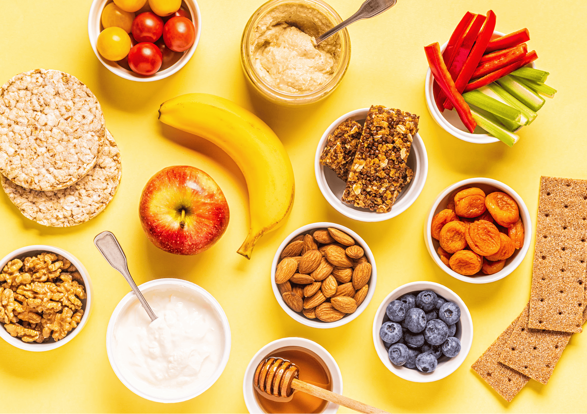 Gesunde Snackauswahl mit Nüssen, Obst, Gemüse, Joghurt und Trockenfrüchten auf gelbem Hintergrund