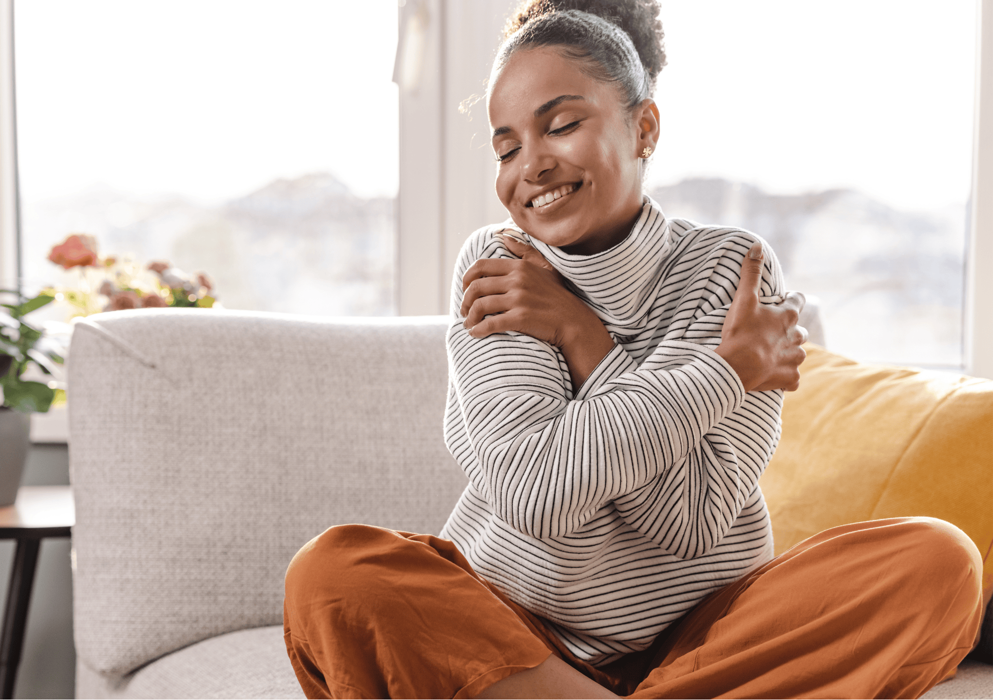 Junge Frau sitzt lächelnd auf dem Sofa und umarmt sich selbst – Symbol für Selbstliebe und Achtsamkeit