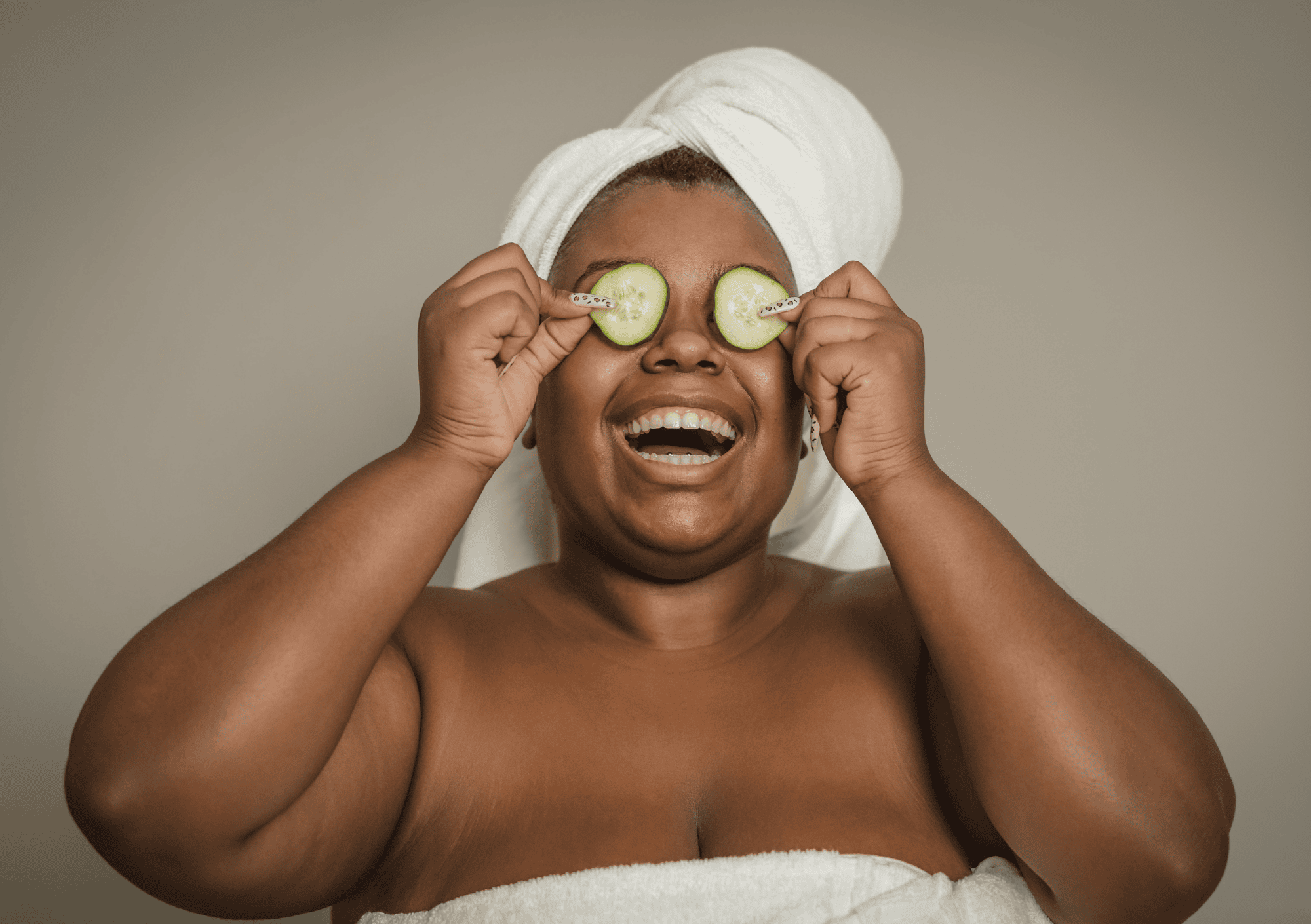Lachende Frau mit Handtuch und Gurkenscheiben auf den Augen beim Wellnessritual
